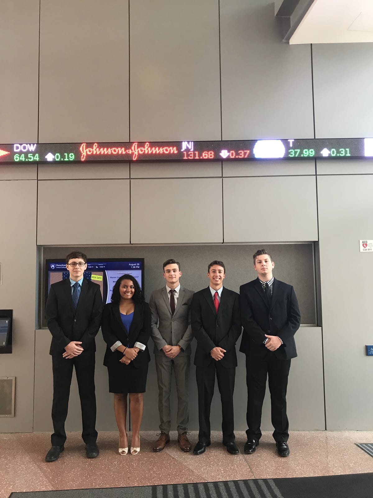 Five members of the PSU Trading Society executive board in business attire beneath the stock ticker in the Business Building lobby
